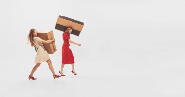Two stylish young women carrying big cardboard boxes isolated over white studio background. Black Friday shopping, sales, moving. Concept of retro fashion, style, youth culture, emotions, beauty, ad