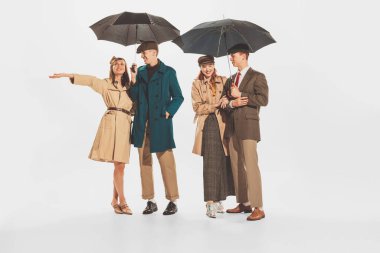 Group of stylish young people in coats and jackets posing with umbrella isolated over white background. Autumn, fall fashion collection. Concept of retro fashion, style, youth culture, beauty, ad