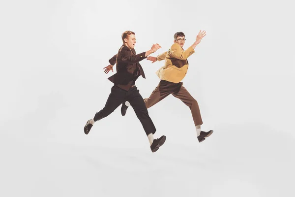 Portrait of two stylish men, friends spending time together, posing in a jump isolated over white studio background. Concept of retro fashion, style, youth culture, emotions, friendship, party, ad
