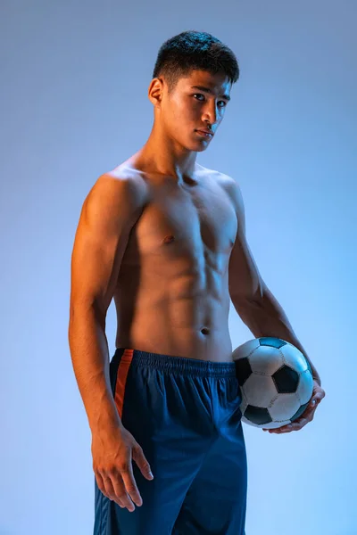 Portrait of young muscular man, football player posing with ball isolated over blue studio background in neon light. Concept of sport, team game, action, motion. Copy space for ad, poster
