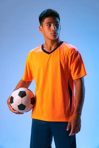 Portrait of young concentrated man, football player posing with ball isolated over blue studio background in neon light. Concept of sport, team game, action, motion. Copy space for ad, poster