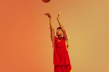 Portrait of young man, basketball player throwing ball into basket isolated over orange studio background in neon light. Concept of healthy lifestyle, professional sport, hobby, power and strength