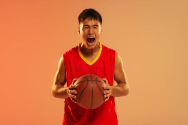 Portrait of young man, basketball player screaming in emotions isolated over orange studio background in neon light. WInning game. Concept of healthy lifestyle, professional sport, hobby, strength