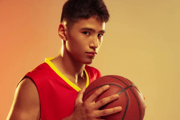 Close-up portrait of young man, basketball player posing with ball isolated over orange studio background in neon light. Concept of healthy lifestyle, professional sport, hobby, power and strength