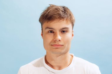 Close-up portrait of young man in white casual T-shirt looking at camera, posing isolated over blue studio background. Concept of emotions, facial expression, lifestyle, fashion, youth