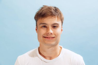 Portrait of young man in casual white T-shirt smiling, posing, looking at camera isolated over blue studio background. Concept of emotions, facial expression, lifestyle, fashion, youth