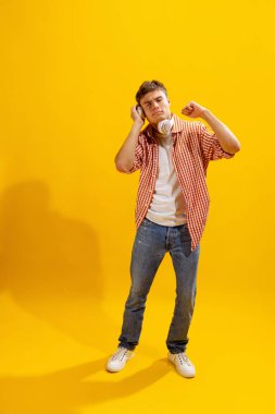 Portrait of young man in checkered shirt posing, listening to music in headphones isolated over yellow studio background. Concept of emotions, facial expression, lifestyle, fashion, youth