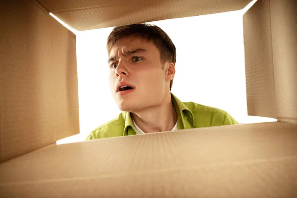 Portrait of young man looking into open cardboard box. Looking with shock and interest. Surprising gift. Concept of emotions, facial expression, lifestyle, fashion, youth