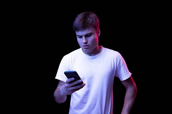 Portrait of young man in white T-shirt posing, reading text message on phone isolated over black background in neon light. Concept of emotions, facial expression, lifestyle, fashion, youth