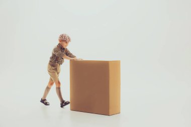 Portrait of child, boy in stylosh ca and cheskered shirt pushing cardboard box isolated over grey studio background. Concept of childhood, friendship, fun, lifestyle, fashion, retro style