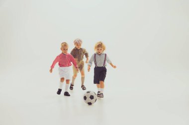 Portrait of little boys, children in vintage clothes playing football together isolated over grey studio background. Concept of childhood, friendship, fun, lifestyle, fashion, retro style