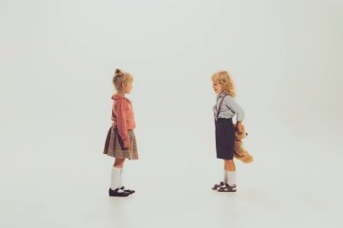 Portrait of two stylish kids posing isolated over grey studio background. Boy hiding behind his back bear toy. Concept of childhood, friendship, fun, lifestyle, fashion, retro style