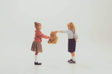 Portrait of two stylish children posing isolated over grey studio background. Boy giving to girl bear toy. Present. Concept of childhood, friendship, fun, lifestyle, fashion, retro style
