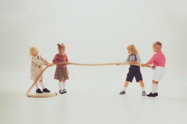 Portrait of children, boys and girls playing together, pulling the rope isolated over grey studio background. Concept of childhood, friendship, fun, lifestyle, fashion, retro style