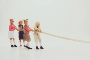 Portrait of children, boys and girls playing together, pulling the rope isolated over grey studio background. Concept of childhood, friendship, fun, lifestyle, fashion, retro style