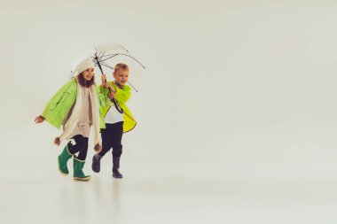 Portrait of boy and girl, children in autumn outfit walking with umbrella isolated over grey studio background. Concept of childhood, friendship, fun, lifestyle, fashion, retro style