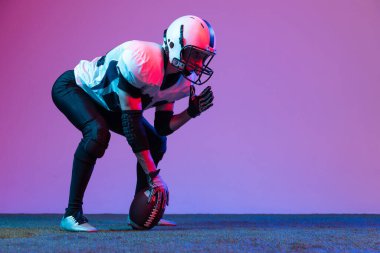 Portrait of concentrated man, professional american football player starting game, posing isolated over purple background in neon light. Concept of active life, team game, energy, sport, competition.