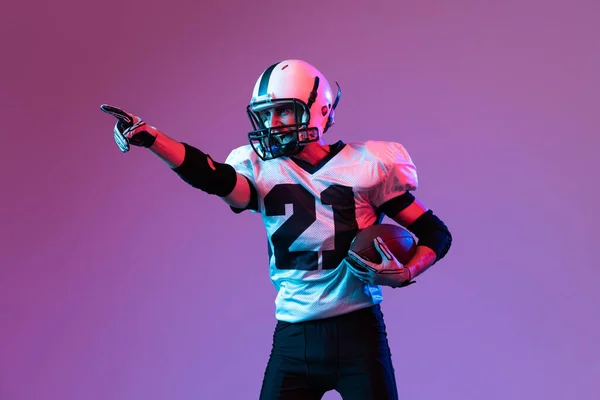 Portrait of emotive man, american football player posing isolated on purple background neon light. Pointing somewhere. Concept of active life, team game, energy, sport, competition. Copy space for ad