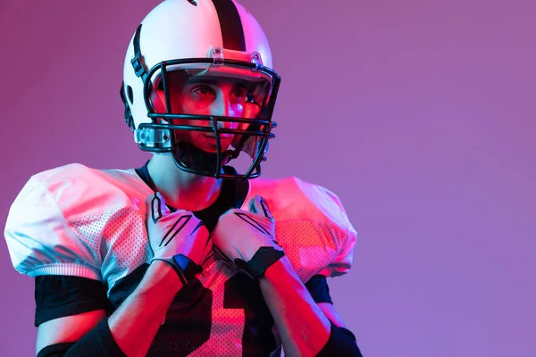 Close-up portrait of young man, american football player in helmet posing isolated over purple background neon light. Concept of active life, team game, energy, sport, competition. Copy space for ad