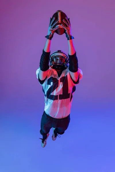 Top view. Portrait of sportive man, american football player in uniform training, catching ball in a jump isolated on purple background in neon light. Concept of team game, energy, sport, competition.