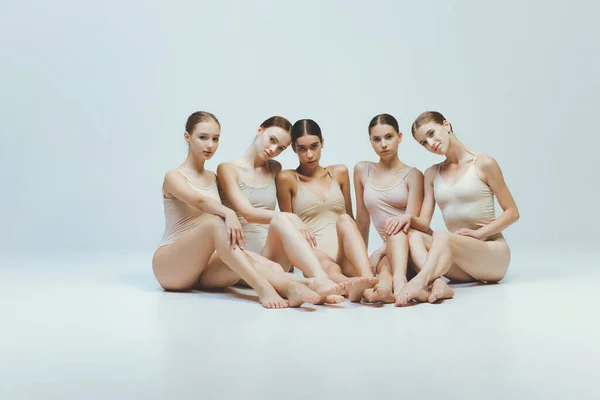 Group of young girls, ballet dancers in beige bodysuits sitting together, posing isolated over grey studio background. Concept of art, beauty, aspiration, creativity, classic dance style, elegance