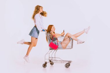 Portrait of stylish young girls going shopping together. Girl sitting on shopping trolley. Cheerful leisure time. Big sales. Concept of youth, retro style, 90s era, fashion, lifestyle, ad