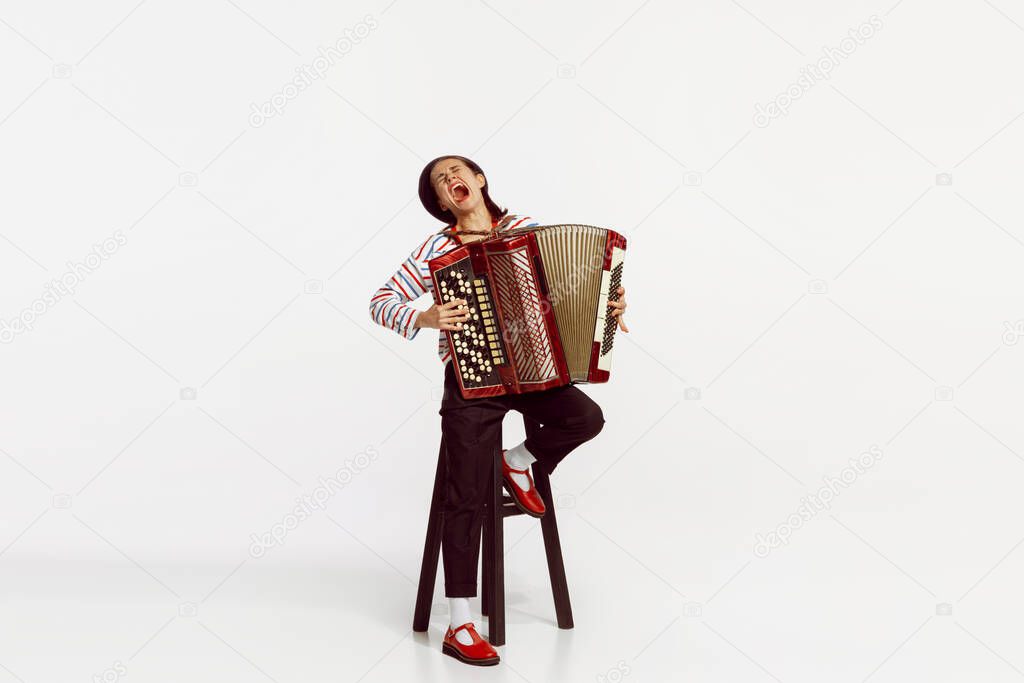 Portrait of talented beautiful woman playing accordion and expressively singing isolated over white studio background. Concept of live music, performance, retro style, creativity, artistic lifestyle