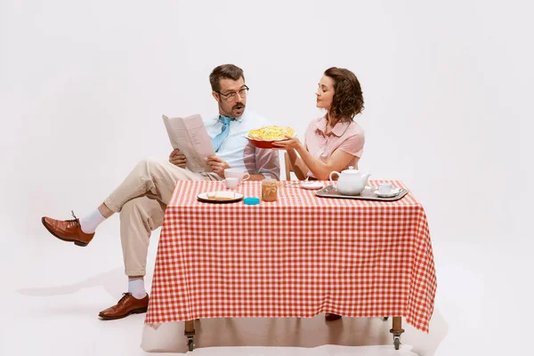 Portrait of loving couple sitting at the table, having breakfast isolated on white background. Wife giving apple pie. Concept of love, relationship, retro style, creativity, family. Copy space for ad