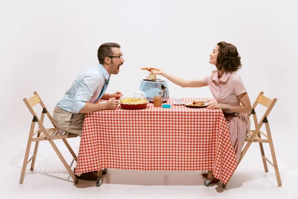 Portrait of couple sitting at the table, having breakfast, wife feeding husband isolated on white background. Toasts for breakfast. Concept of love, relationship, retro style, creativity, family.