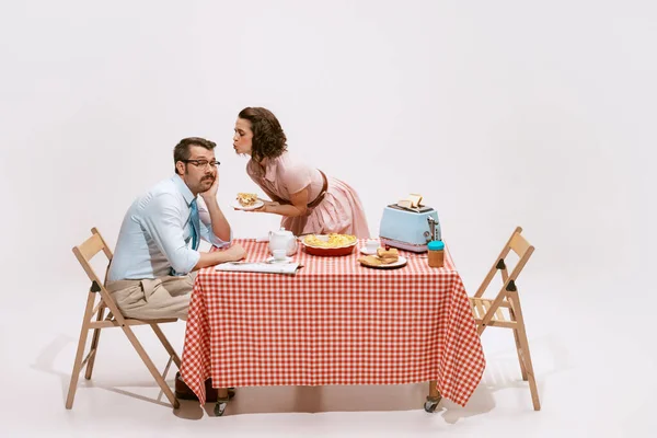 Portrait of couple sitting at the table, having breakfast isolated on white background. Sleepy man and caring woman. Concept of love, relationship, retro style, creativity, family. Copy space for ad