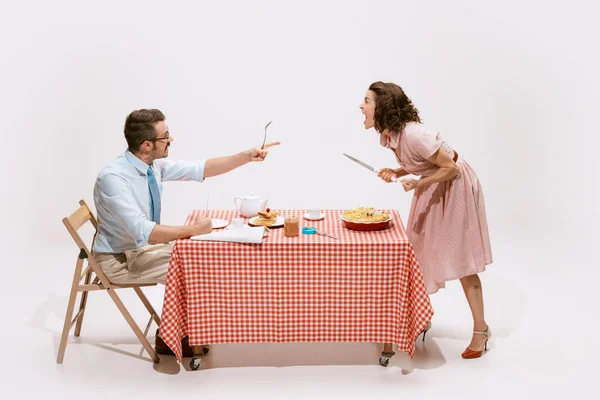 Portrait of loving couple sitting at the table, having breakfast and quarrelling isolated on white background. Concept of love, relationship, retro style, creativity, family. Copy space for ad