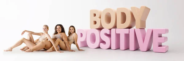 Three young women with different body shapes sitting on floor in underwear near big 3D lettering of body positive. Natural beauty. Concept of skincare, cosmetology, spa, plastic surgery, wellness