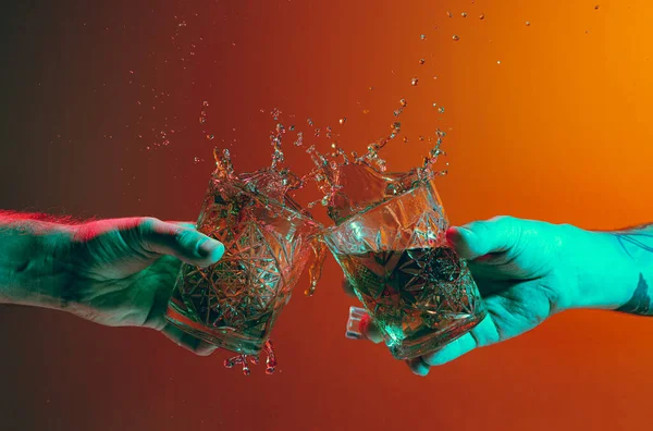 Male hands clinking glasses with whiskey over dark orange background in neon light. Hard drink. Concept of alcoholic drinks, party, taste. Food and drink art. Copy space for ad