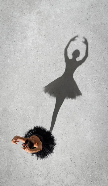 Portrait of young ballerina in black dress performing, standing on pointe isolated on grey wall background. Top view. Shadow element. Concept of classic ballet, inspiration, beauty, dance, creativity