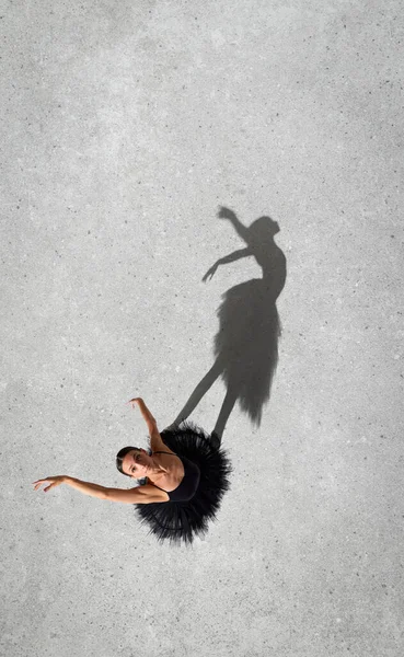 Portrait of young ballerina in black dress performing, looking upwards isolated over grey wall background. Top view. Shadow element. Concept of classic ballet, inspiration, beauty, dance, creativity