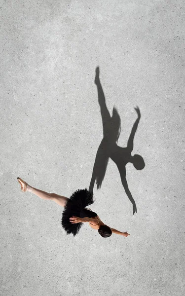 Portrait of young ballerina in black dress performing isolated over grey wall background. Top view. Shadow element. Twine. Concept of classic ballet, inspiration, beauty, dance, creativity