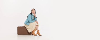 Kaliteli bir bavul üzerinde oturan genç kadın beyaz arka planda izole görünüyor. Seyahat ediyorum. Geçmişe dönük moda, tarz, macera, yaratıcılık, gençlik kültürü, duygular, reklam