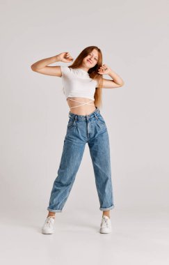 Gri stüdyo arka planında günlük kıyafetler, kot pantolon ve beyaz bluzla poz veren kızıl saçlı güzel bir kızın tam boy portresi. Duyguların kavramı, yaşam tarzı, yüz ifadesi, güzellik