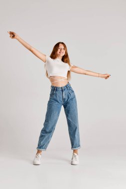 Gri arka planda izole edilmiş mutlu duygularla günlük kıyafetlerle poz veren güzel bir genç kızın tam boy portresi. Duyguların kavramı, yaşam tarzı, yüz ifadesi, güzellik, moda, gençlik