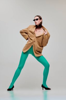 Yeşil taytlı, güneş gözlüklü ve gri arka planda izole edilmiş büyük beden ceketli güzel bir genç kadının portresi. Modern moda kavramı, sanat fotoğrafçılığı, stil, nonoş, güzellik, reklam