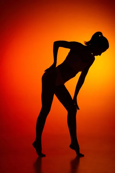 Silhouette of female full-length body isolated over orange background. Side view. Woman leaning with hand on her knee. Body art, aesthetics. Concept of beauty, femininity, health, sport, lifestyle