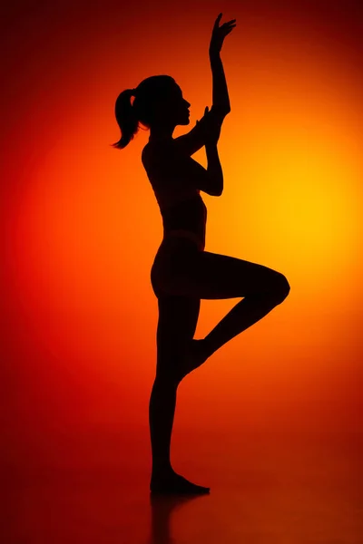 Silhouette of female full-length body isolated over orange background. Standing on one leg. Body art, aesthetics. Concept of beauty, femininity, health, sport, lifestyle