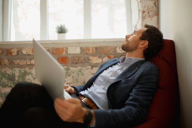 Düşünceli bir ifadeyle genç iş adamının portresi dizüstü bilgisayarla yerde oturuyor ve çalışıyor, yeni projeler düşünüyor. İş kavramı, zorluklar, iş, meslek