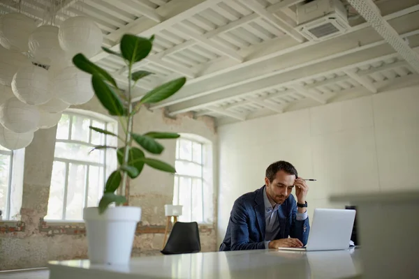 Portrait of young businessman sitting in empty building at the table, looking in laptop with thoughtful expression. Going out of business. Crisis. Concept of business, challenges, work, occupation