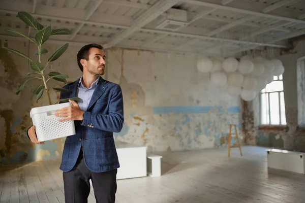 Portrait of young businessman walking out empty room with flower. Business closure. Depression and crisis. Concept of business, challenges, work, occupation, failure, bankruptcy, liquidation