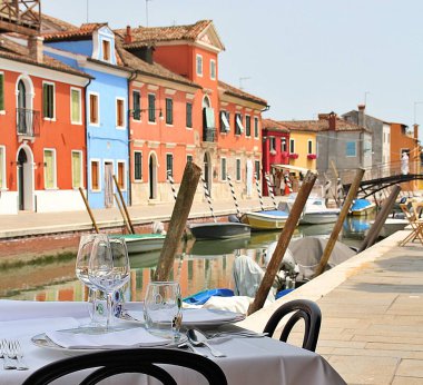 Restoran masası, restoran masası Burano 'daki kanalın kıyısındaki caddeye açılacak, dışarıda restoran masaları, beyaz masa örtüsü, çatal bıçak takımı, tabak ve bardaklar, su kanalının arka planında bulanık renkli evler.