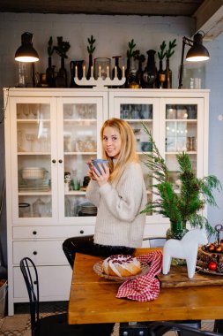 Yeni yıl süslemeleri için mutfakta çay içen güzel sarışın bir kadın..