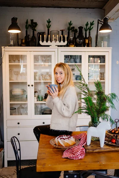 Yeni yıl süslemeleri için mutfakta çay içen güzel sarışın bir kadın..