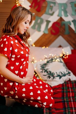 Güzel hamile kadın kırmızı pijamalarıyla yatak odasında, Noel ağacının yanında..