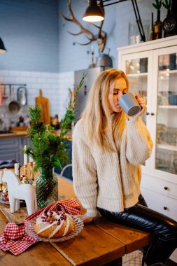 Yeni yıl süslemeleri için mutfakta çay içen güzel sarışın bir kadın..